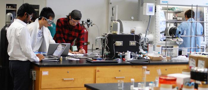 Students work with a professor, surrounded by chemistry equipment.